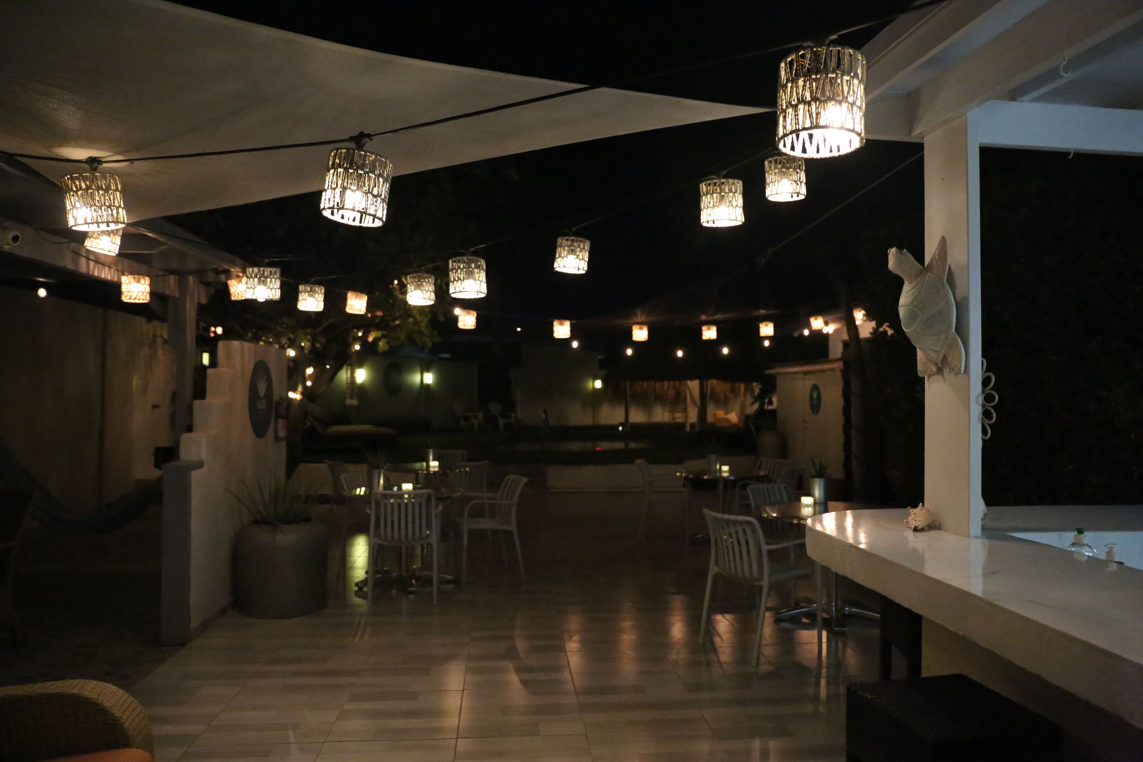 An outdoor seating area with tables and chairs, overhead lighting, and a statue on the right.