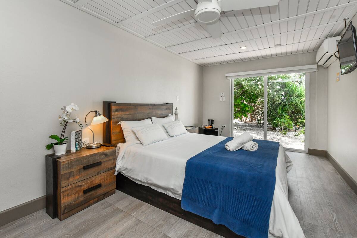 A modern bedroom with a large bed, wooden nightstands, a blue blanket, and a view of greenery through the window.