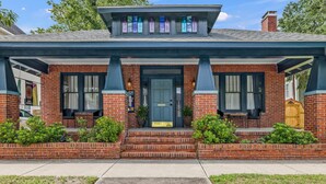 A two-story brick house with a dark blue roof, a front porch with steps, and a small garden area.