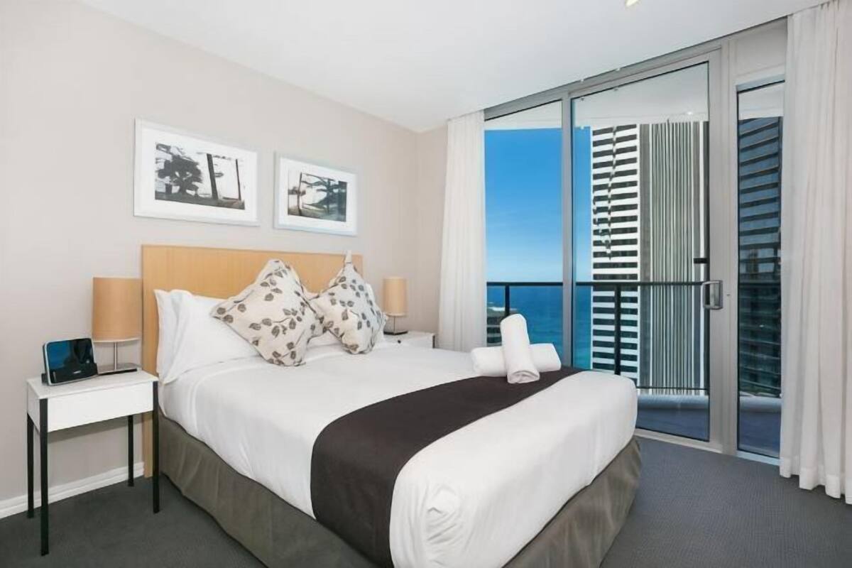 A hotel room with a bed, a nightstand, a laptop, a window with blinds, and a balcony with a view.