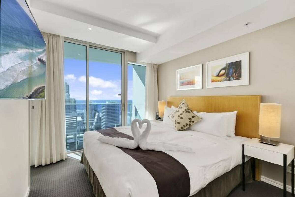 A hotel room with a large bed, a nightstand, a lamp, a painting of a cityscape, and a view of the city through the window.