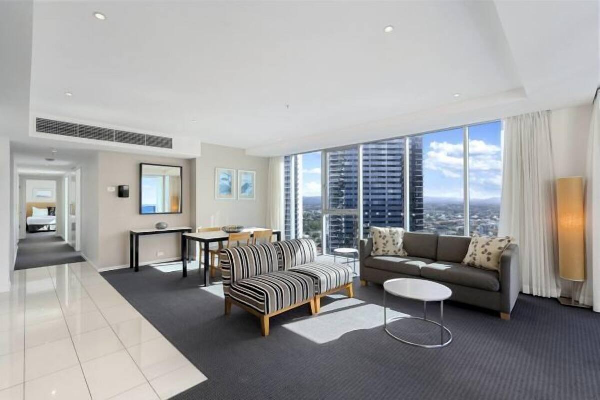 A modern hotel room with a large window, a sofa, a dining area, and a flat-screen TV.