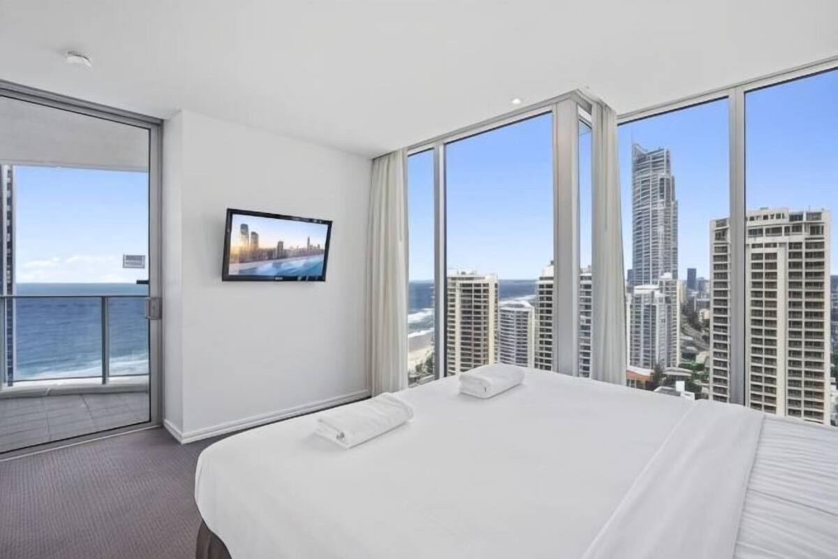A hotel room with a large bed, a television on the wall, and a view of the city and ocean.