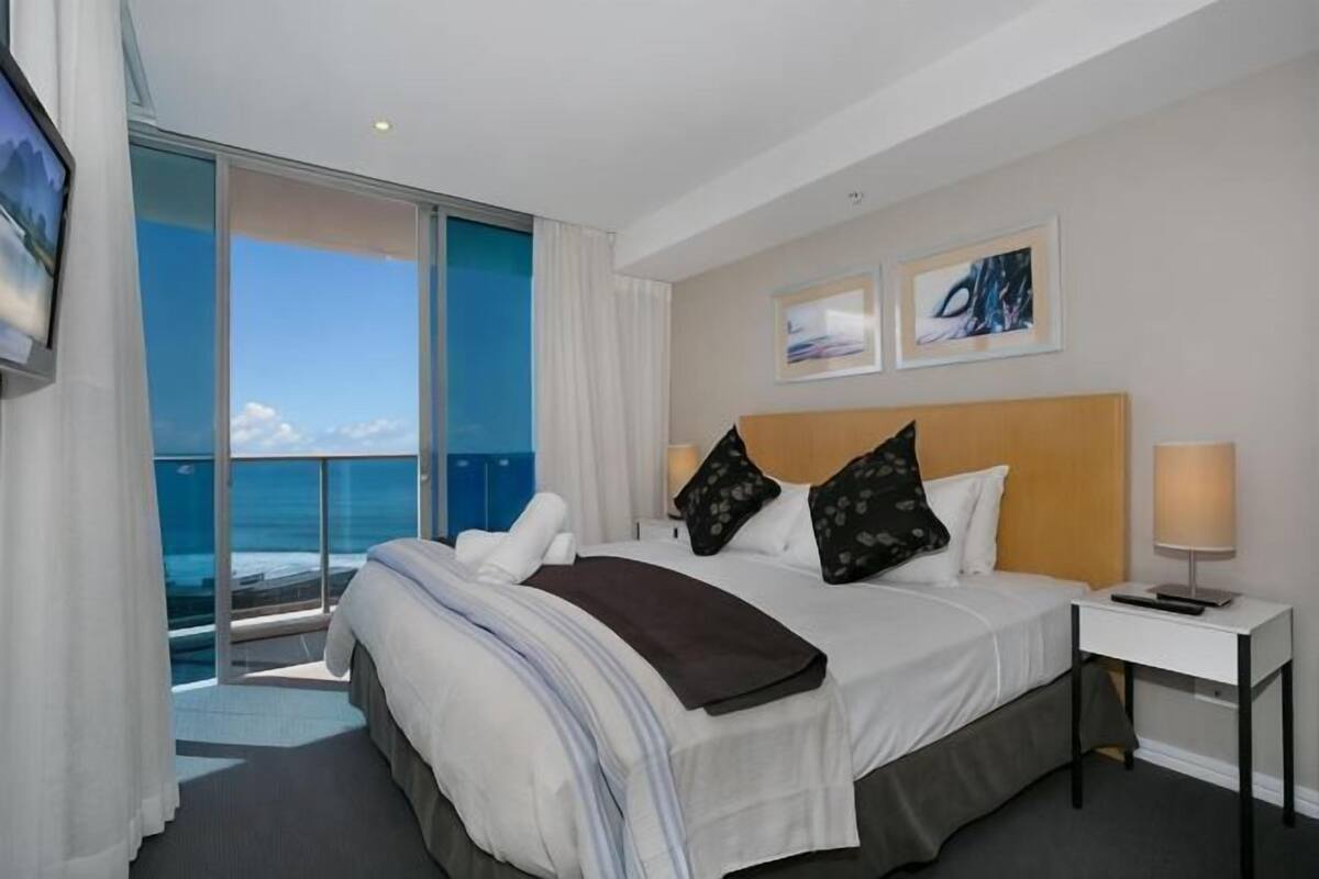A hotel room with a bed, a bedside table, a TV, and a view of the ocean.