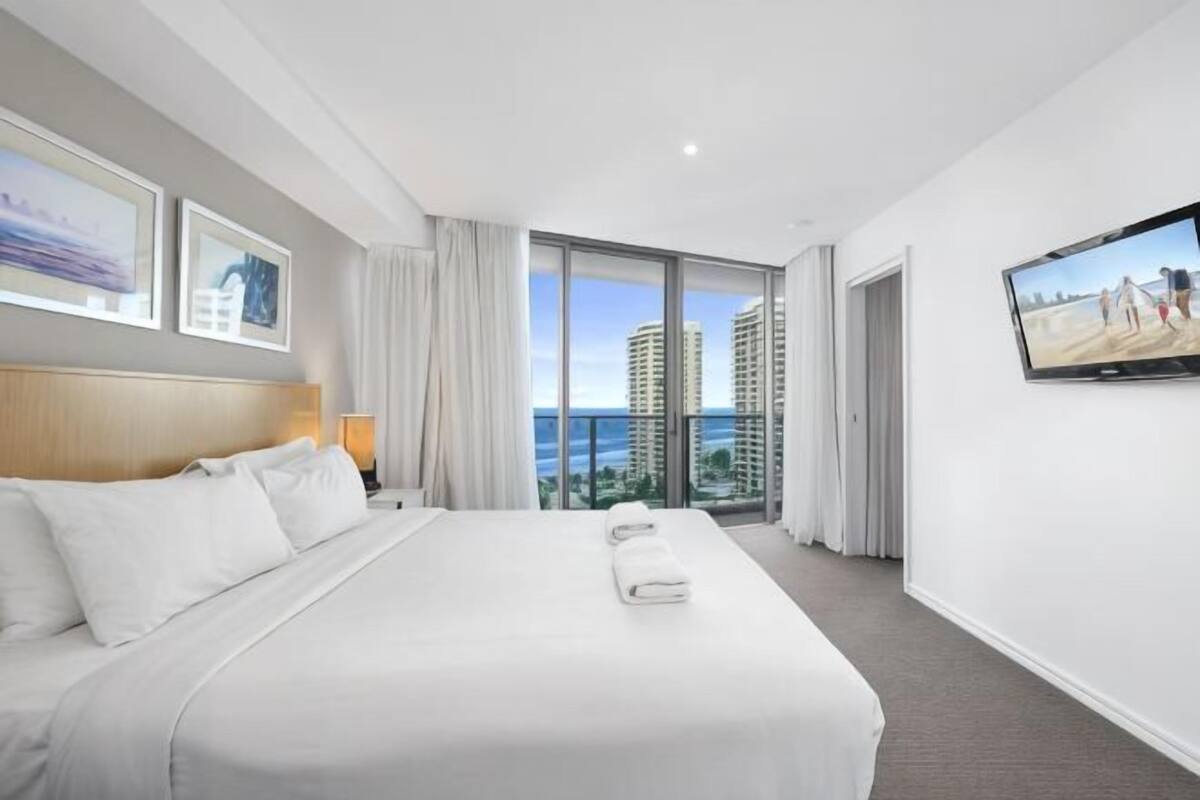 A hotel room with a large bed, a TV, a balcony with a view of the city, and a window with curtains.