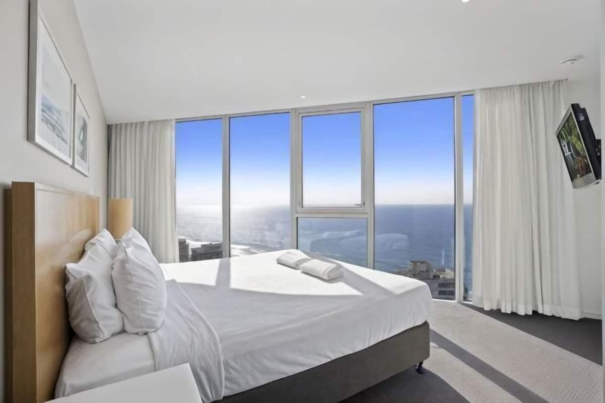 A hotel room with a large bed, a TV mounted on the wall, and a city view through large windows.