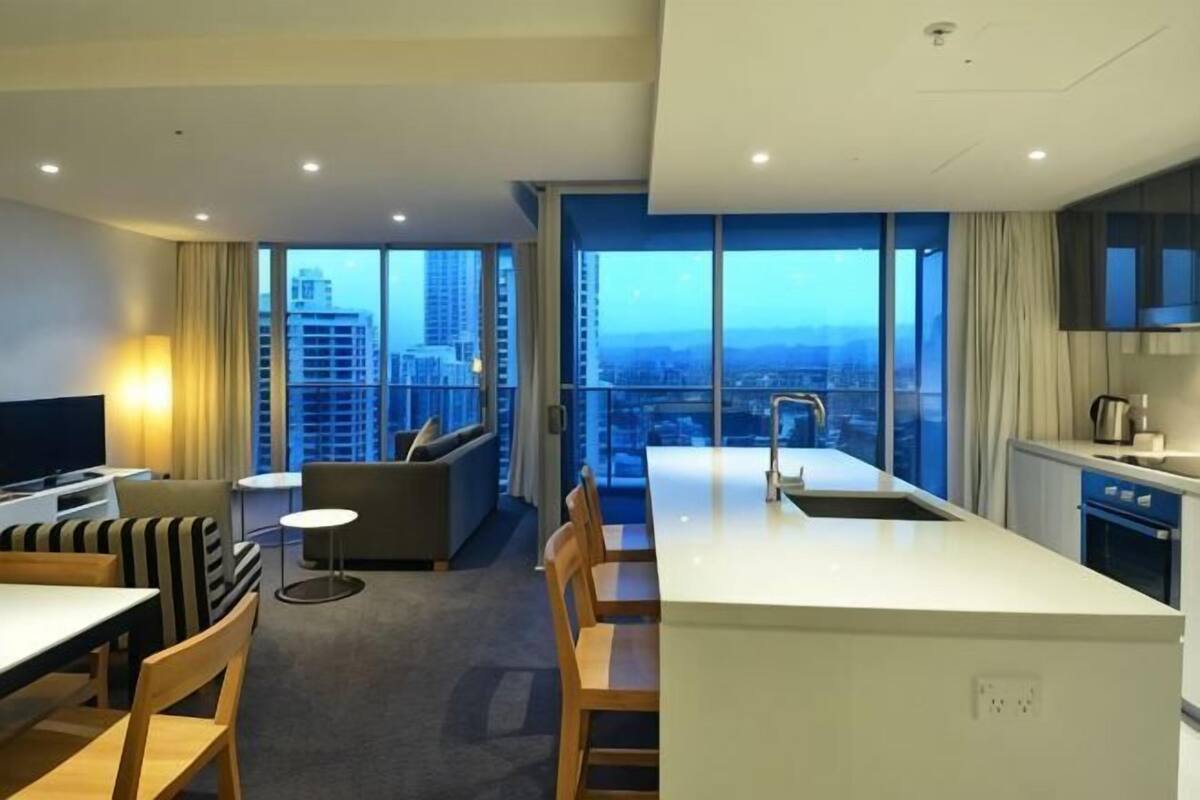 A modern hotel room with a kitchen, dining area, and a large window offering a city view.