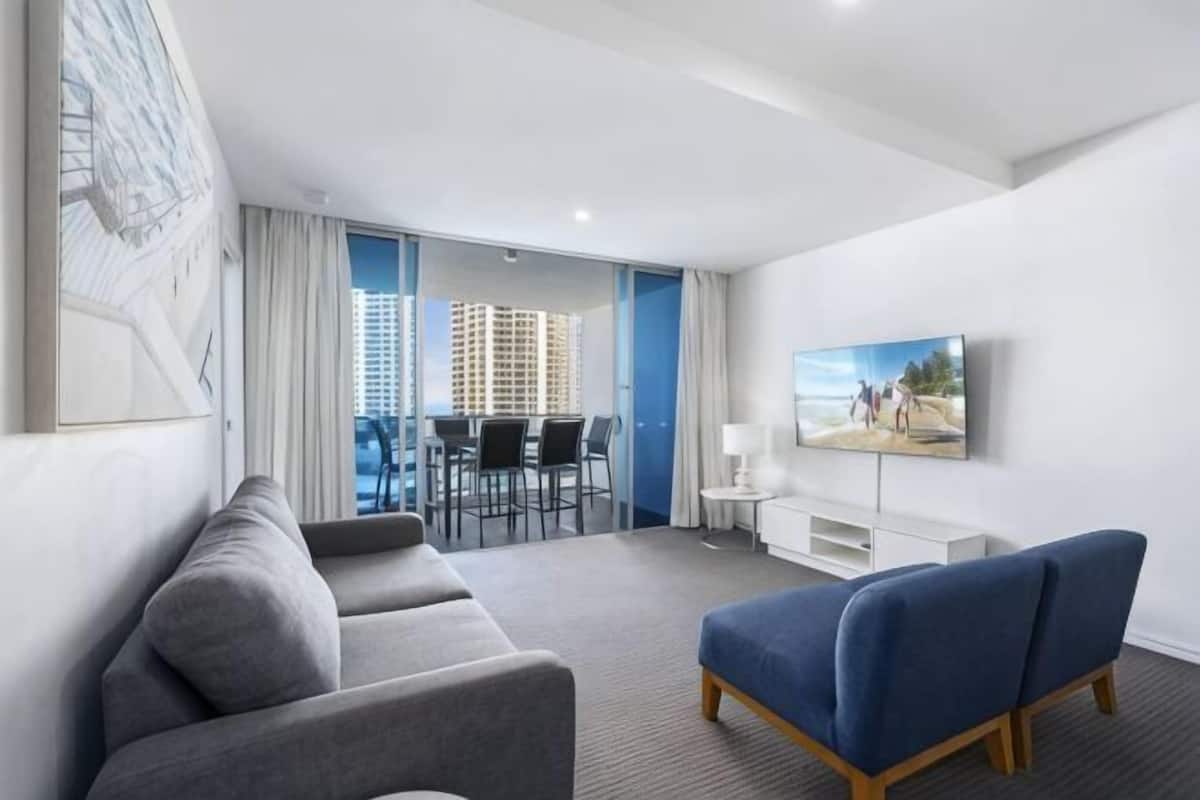 A modern hotel room with a grey sofa, a blue armchair, a flat-screen TV, a dining area with a table and chairs, and a balcony with a view of the city.