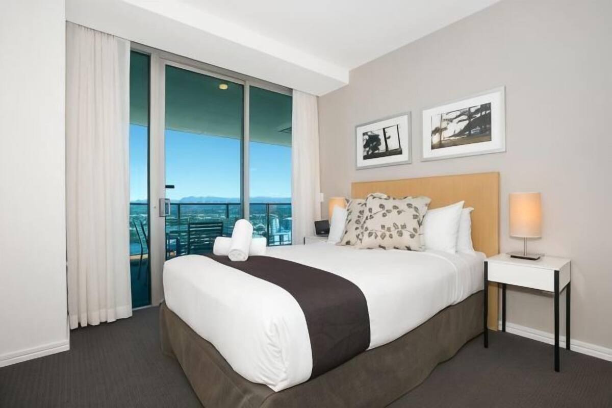 A hotel room with a large bed, a nightstand, a lamp, and a city view through a sliding door.