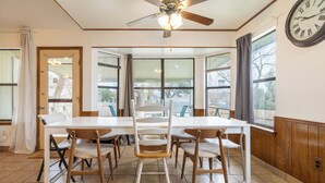 Comedor con una mesa blanca, seis sillas de madera, un ventilador de techo y ventanas grandes con cortinas.