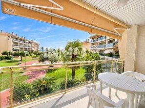 A balcony with a glass railing, a white table, and chairs. There is a view of a landscaped area with palm trees and a building in the background. - Golf Sant Jordi - La Llosa, sea views, parking  (Cambrils)