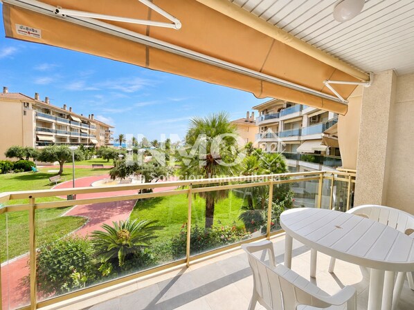 A balcony with a glass railing, a white table, and chairs. There is a view of a landscaped area with palm trees and a building in the background.