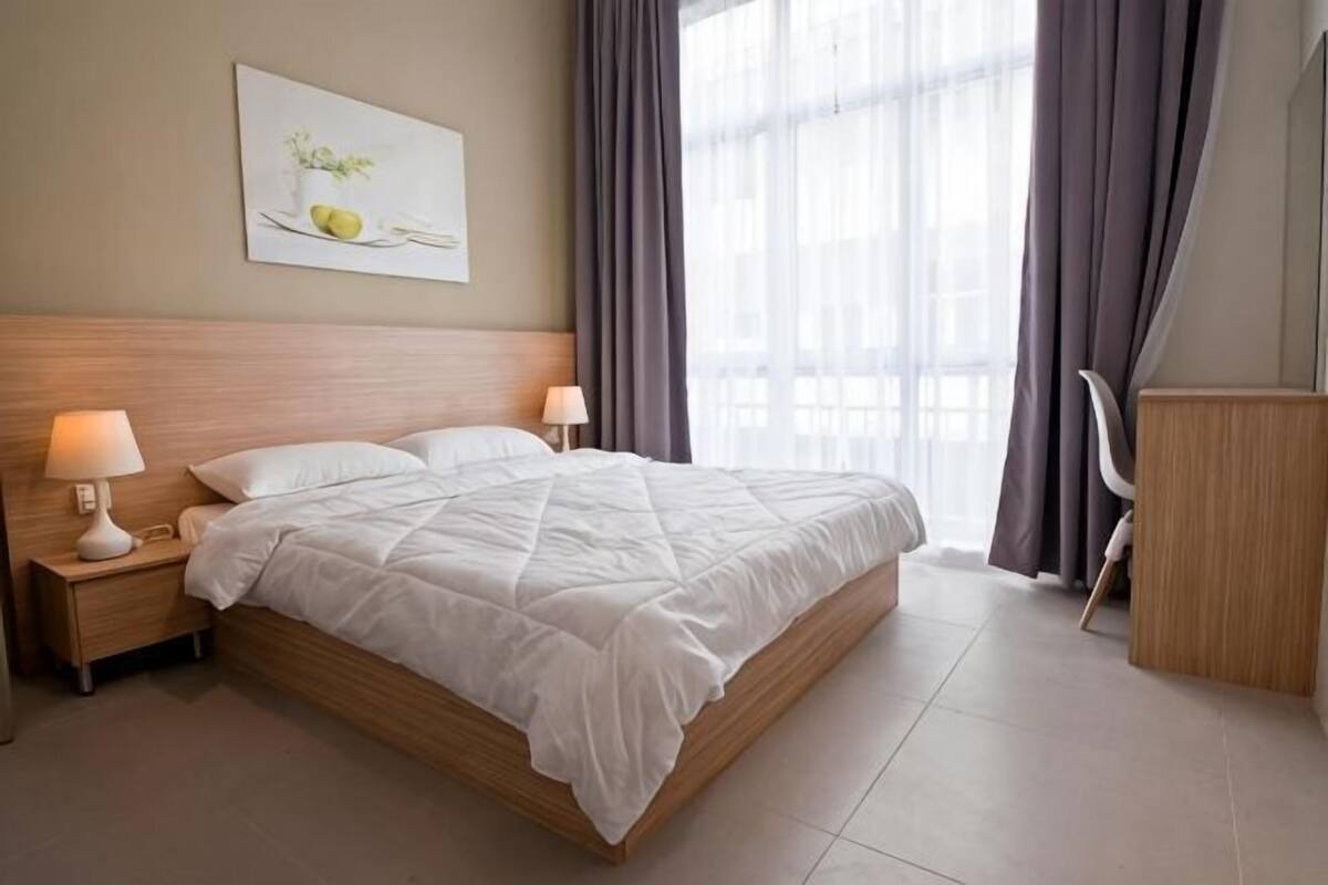 A neatly made bed with white bedding, a wooden headboard, two bedside lamps, a nightstand, a chair, and a window with curtains.