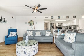 A living room with a ceiling fan, a blue armchair, a grey sofa, and a round coffee table. - Holiday Villas III - 410 Stunning Condo Intracoastal View Balcony , heated pool! (Indian Rocks Beach)