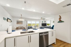 A modern kitchen with a central island, a dining area with a white table and chairs, and a living room with a sofa and TV.