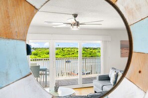 A room with a ceiling fan, a balcony with a view of greenery, and a seating area with a table.