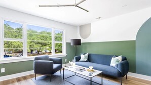 A modern living room with a sofa, armchair, and coffee table. There is a large window with a view of trees and a road. - The Double Up | The Quad in Park Circle (North Charleston)