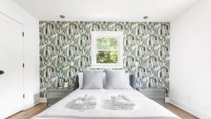 A modern bedroom with a geometric-patterned wallpaper, a neatly made bed with two pillows, and a window with a view of greenery. - The Double Up | The Quad in Park Circle (North Charleston)