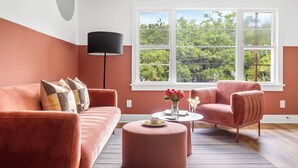 A modern living room with a sofa, armchair, and ottoman, a round coffee table with a vase of flowers, and a large window with a view of trees. - The Duo at the Quad in Park Circle (North Charleston)