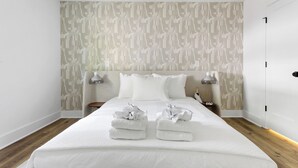 A modern hotel room with a large bed, white linens, and a patterned wallpaper. There are two bedside tables with lamps and a small nightstand with a lamp. - The Duo at the Quad in Park Circle (North Charleston)