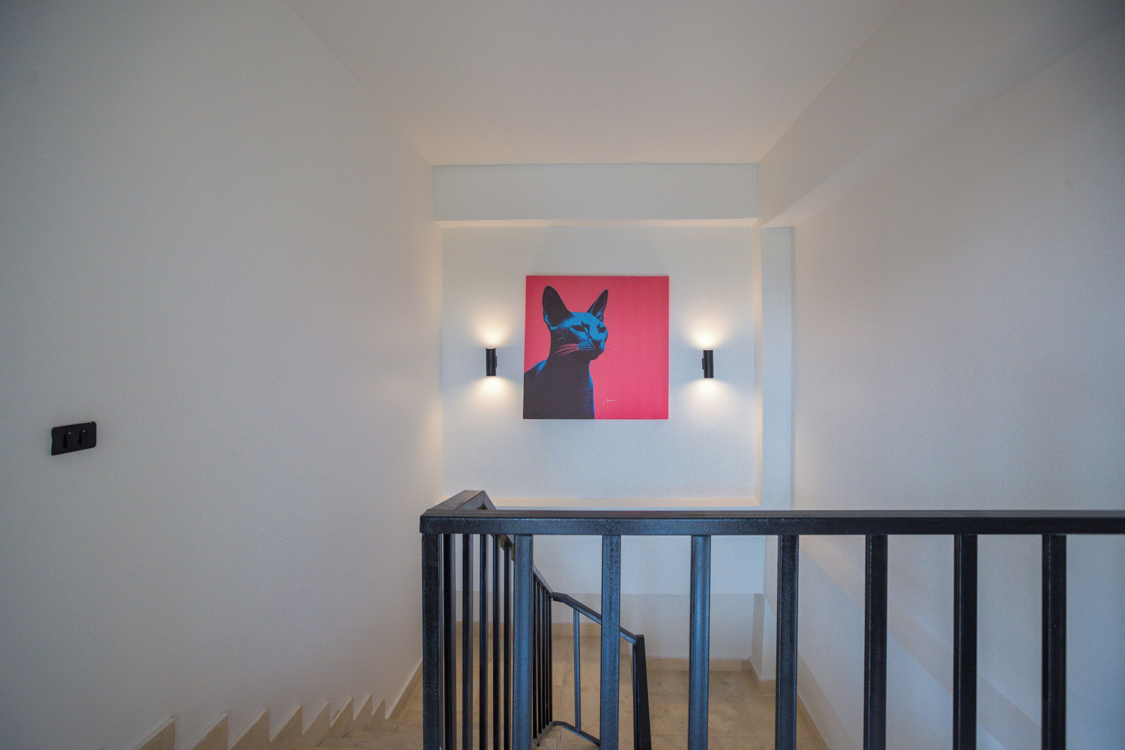 Un escalier avec une rampe noire, un tableau représentant un chat accroché au mur et deux lampes fixées au mur.