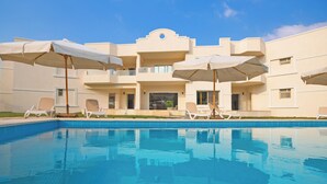 A swimming pool with lounge chairs and umbrellas, a building with balconies, and a clear blue sky. - Seven Stays Zayed (Sheikh Zayed City)