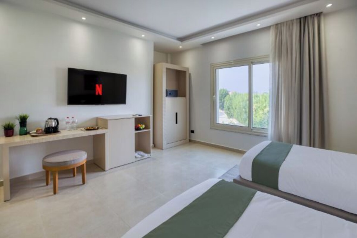 A modern hotel room with a bed, a desk with a chair, a TV mounted on the wall, and a window with curtains.