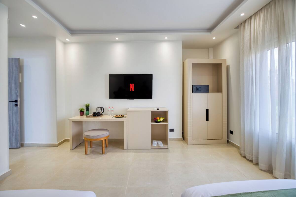 A modern hotel room with a flat-screen TV mounted on the wall, a desk with a chair, a wardrobe, and a bed.