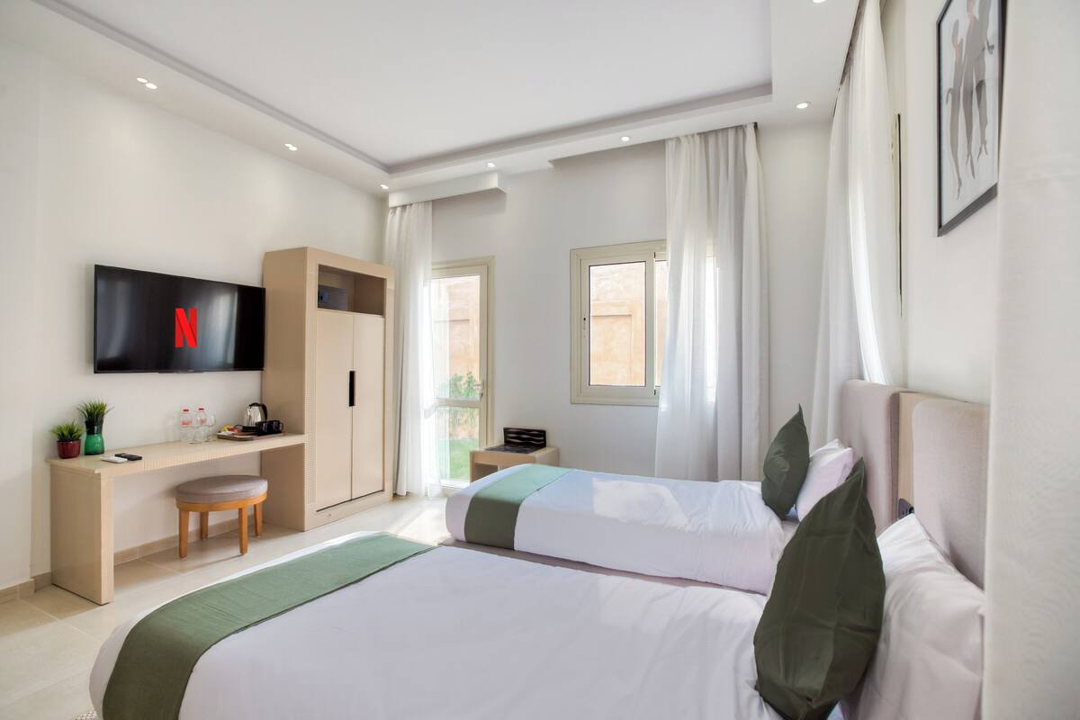 A hotel room with two beds, a desk with a TV, a wardrobe, and a window with curtains.
