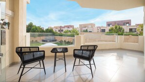 A balcony with two black chairs and a small table, overlooking a residential area with modern buildings and a swimming pool. - Seven Stays Zayed (Sheikh Zayed City)