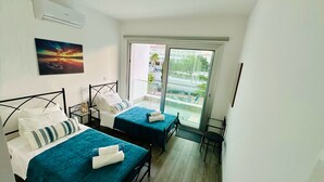 A hotel room with two beds, a small table, a chair, a window with a view of the outside, and a painting on the wall.