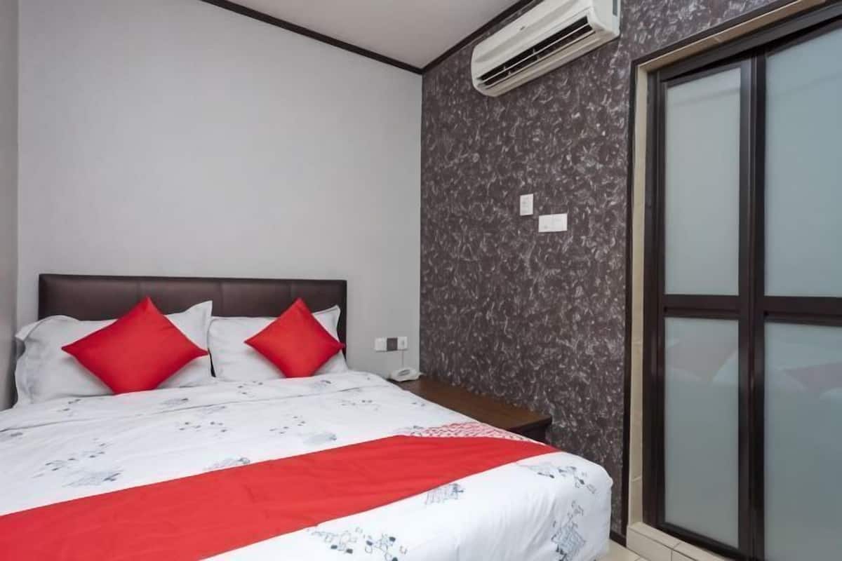 Een slaapkamer met een bed, rode kussens, een wit en rood dekbed en een raam met zonwering, plus een airconditioner aan de muur.