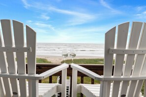 Two white Adirondack chairs facing a beach with a wooden deck and two empty wine glasses on the backrest.