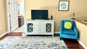 Un salon avec un téléviseur posé sur un meuble blanc, un fauteuil bleu agrémenté d’un coussin coloré et une œuvre d’art encadrée accrochée au mur.