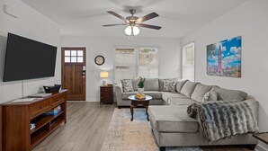 A living room with a flat-screen TV, a ceiling fan, a sofa, a coffee table, and a painting of the Eiffel Tower. - 3BR Walkable Downtown Retreat ~ King Bed + Porch (Salisbury)