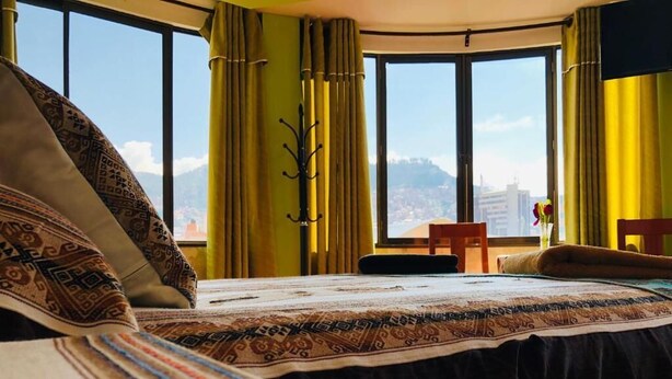 A hotel room with a large bed, a view of the city through large windows, and a chair by the window. - Hostal Iskanwaya (La Paz)