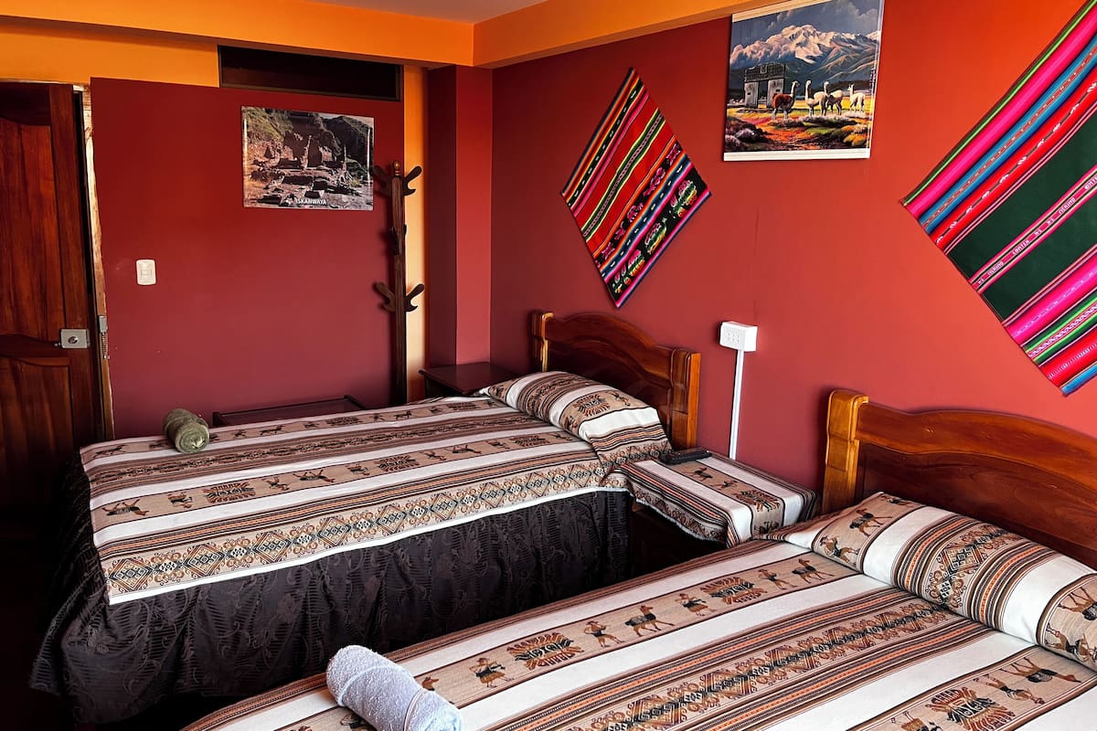 A room with two beds, each with patterned bedding and a wooden headboard. There are two wall-mounted paintings, one depicting a mountainous landscape and the other featuring colorful stripes.