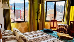 A bedroom with a large window, a bed with patterned bedding, a chair, and a small table with a vase. - Hostal Iskanwaya (La Paz)