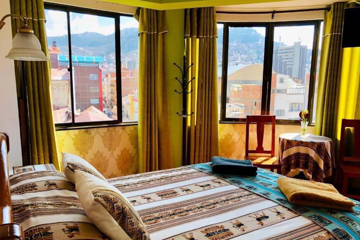 A bedroom with a large window, a bed with patterned bedding, a chair, and a small table with a vase.