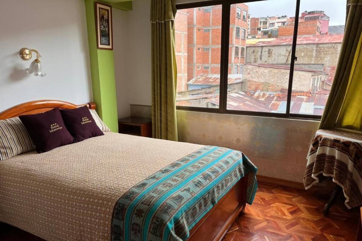 A bedroom with a bed, a window with a view of buildings, a table with a cloth, and a wall-mounted light fixture.
