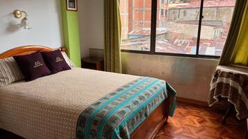 Un dormitorio con cama, ventana con vistas a edificios, una mesa con mantel y una lámpara fijada a la pared.