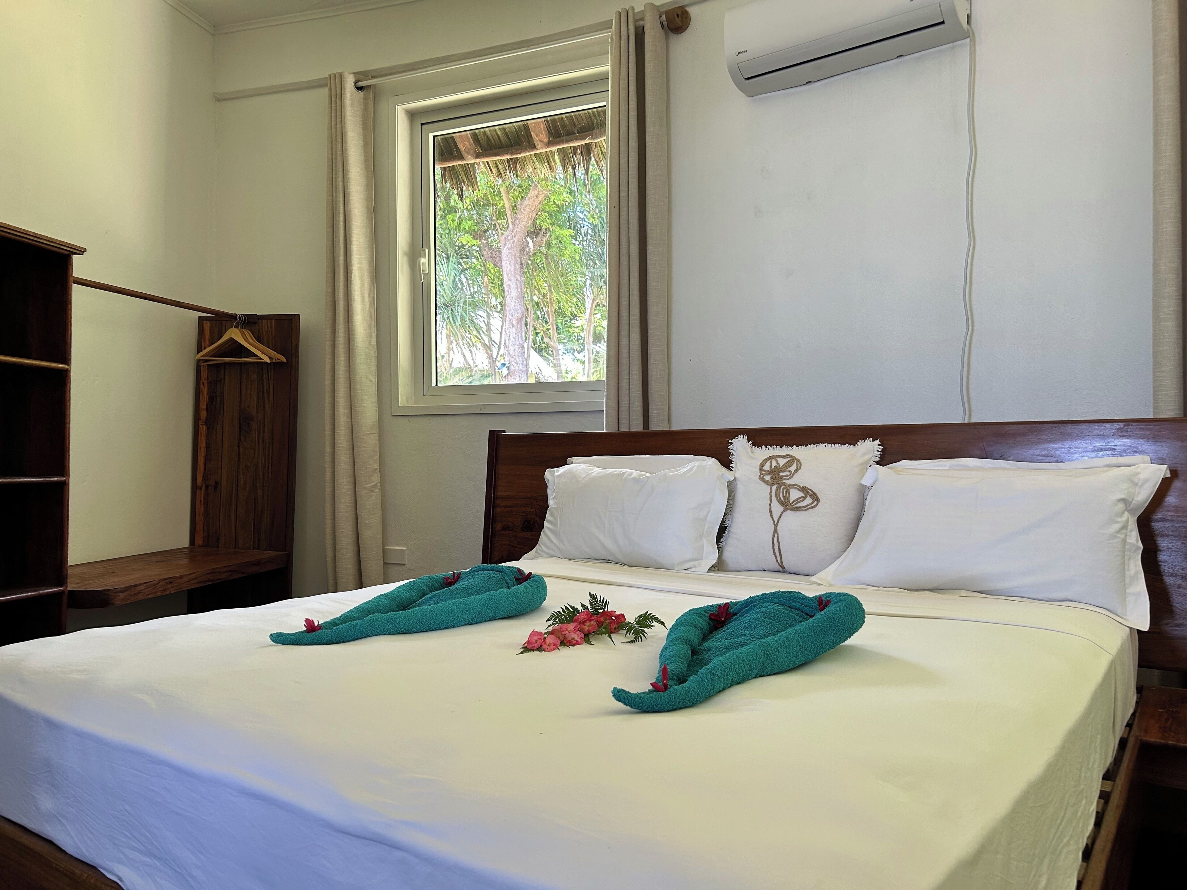 A neatly made bed with white linens and two teal towels, a wooden headboard, a window with a view of trees, and an air conditioning unit on the wall.