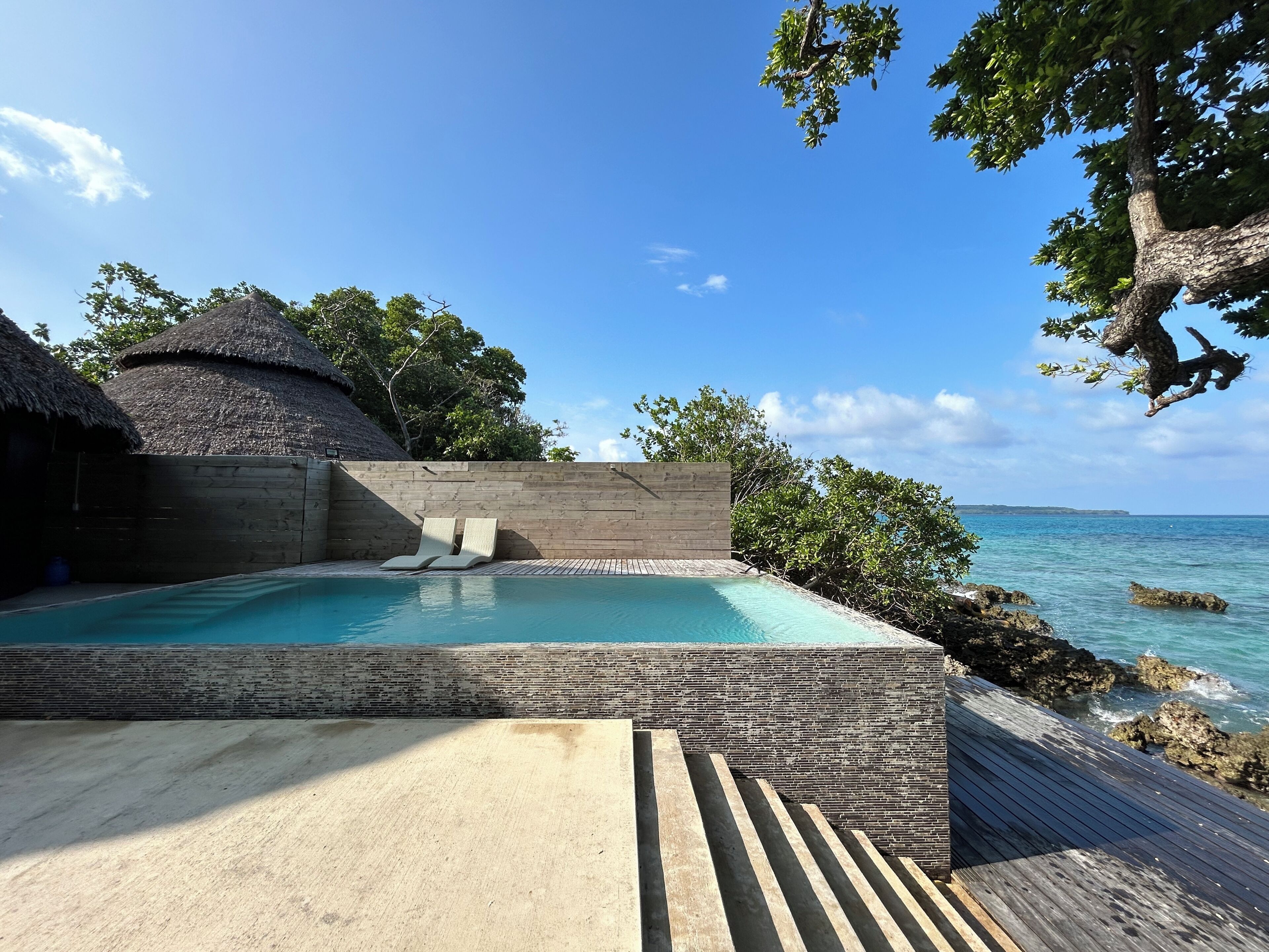 A infinity pool with a wooden deck, a thatched-roof structure, and a view of the ocean.