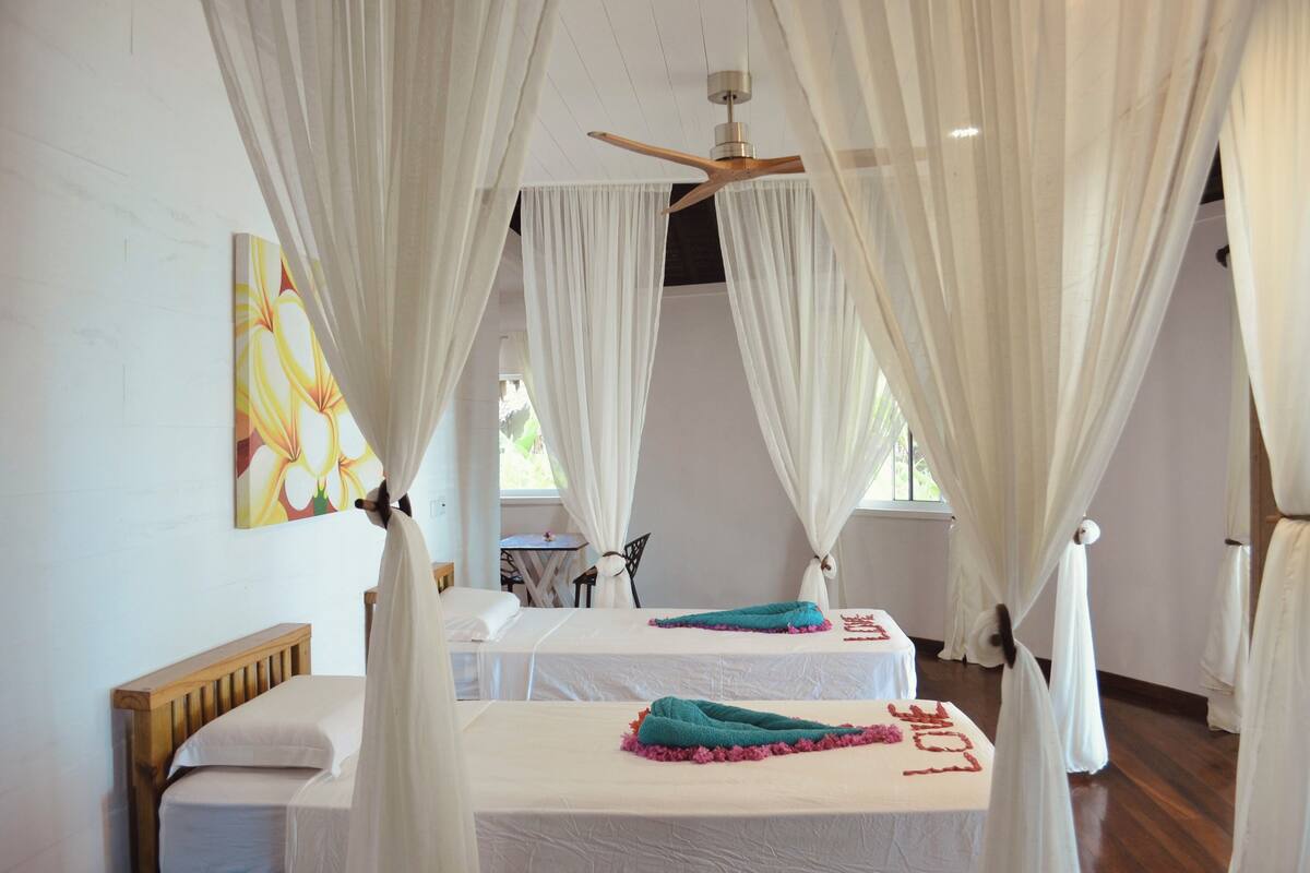 A room with two beds, white curtains, a ceiling fan, and a painting on the wall.