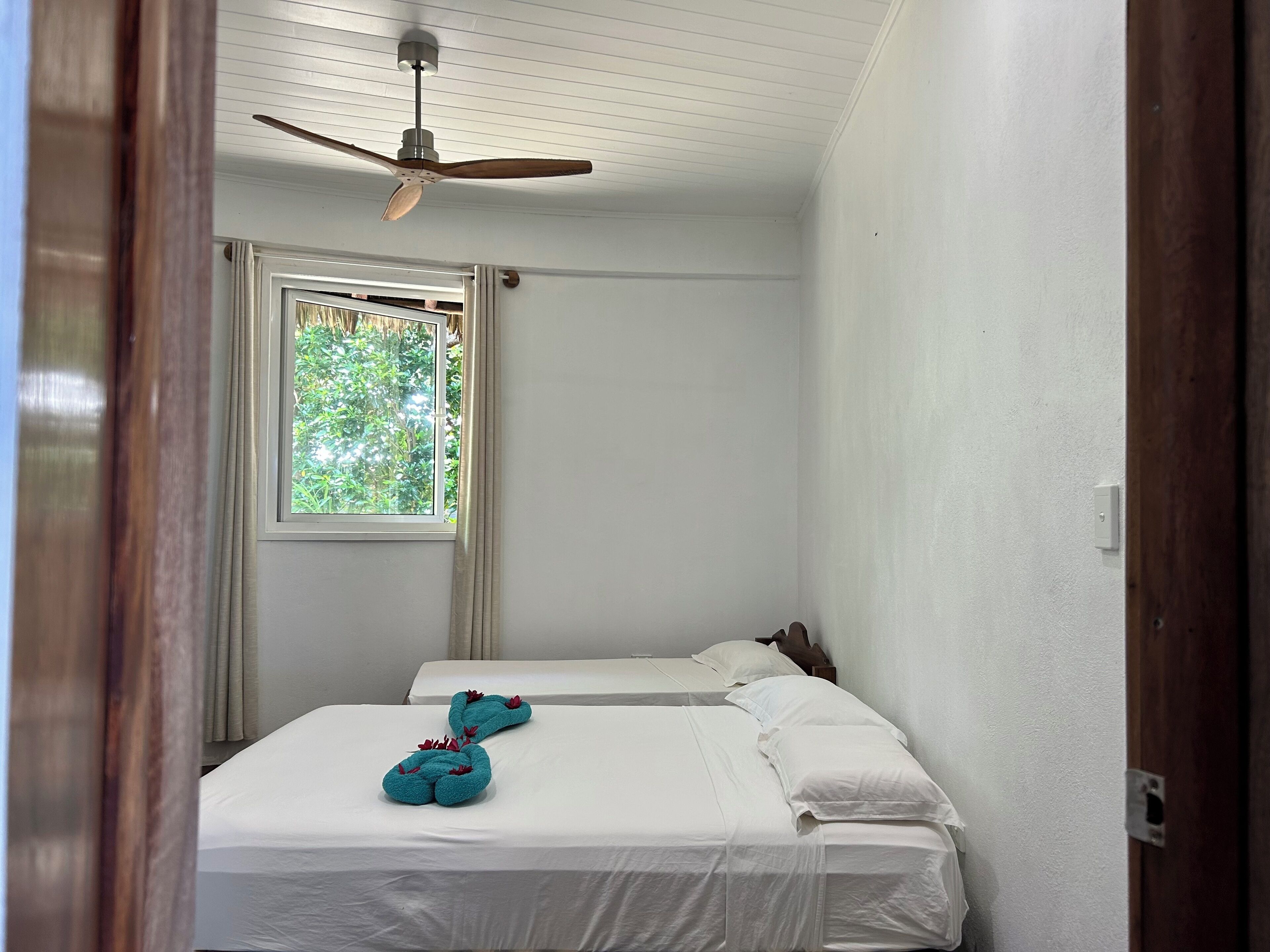 A room with two beds, a ceiling fan, a window with curtains, and a view of greenery outside.