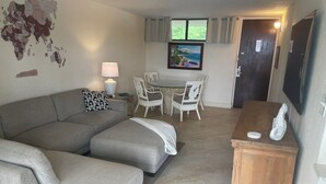 A living room with a grey sectional sofa, a small wooden table with decorative items, a dining area with white chairs, and a painting above the door. - NEW LISTING- Ideal Oceanside Location and Recent Renovation / DREAM Combination! (St Thomas)