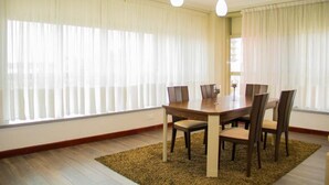Un comedor con una mesa y sillas de madera, ventanas amplias con cortinas ligeras y lámparas colgantes.