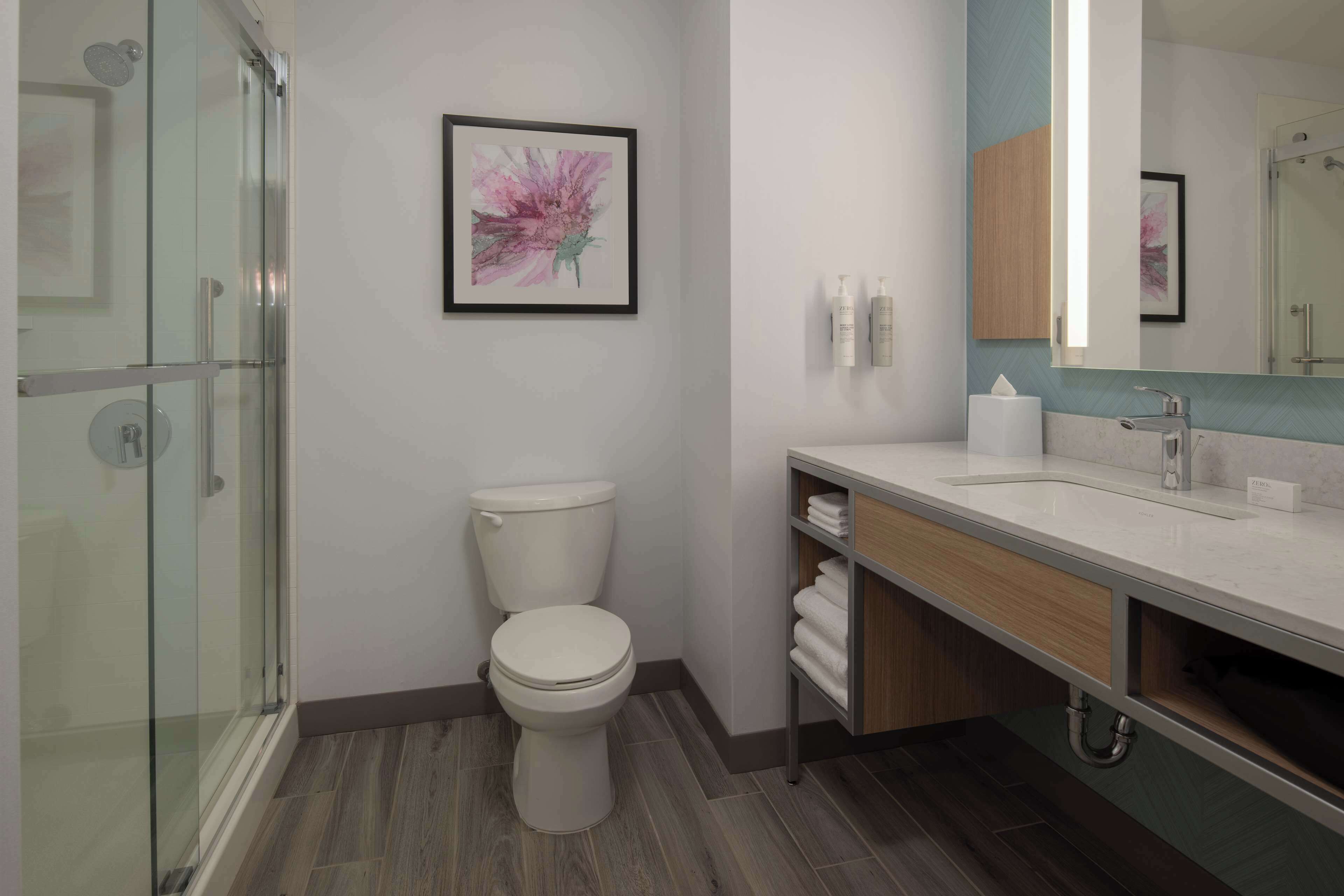 A modern bathroom with a glass shower enclosure, a white toilet, a vanity with a mirror, and a framed floral artwork on the wall.