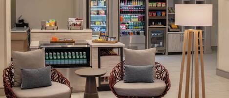 Un moderno vestíbulo de hotel con dos sillones de mimbre, una mesita redonda, una lámpara blanca y un refrigerador lleno de bebidas y aperitivos.