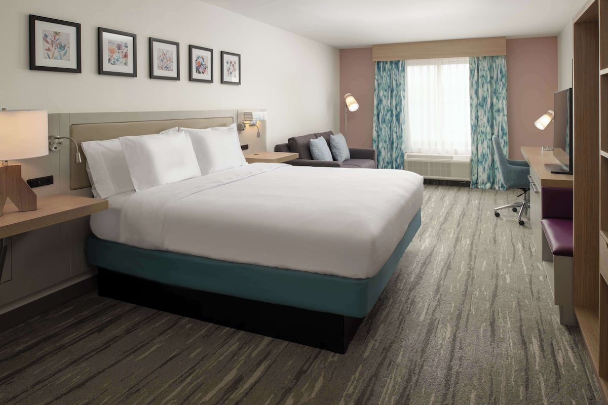 A hotel room with a large bed, a desk, a chair, and a TV. There are framed pictures on the wall and a window with curtains.